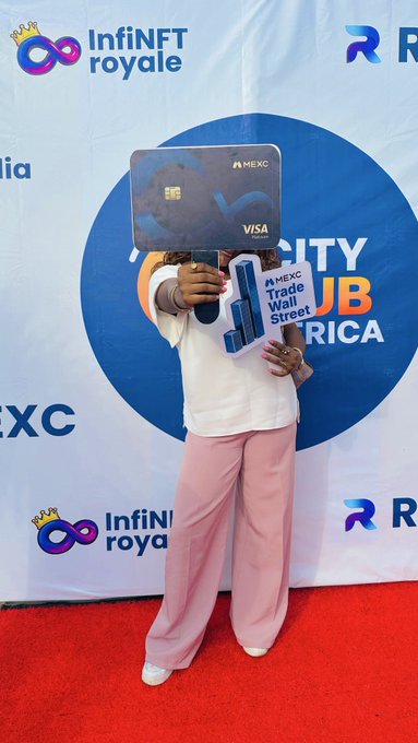 A person on a red carpet at a MEXC event holding up a large MEXC Visa card prop, with 'InfiNFT royale' and 'MEXC Trade Wall Street' branding visible in the background.
