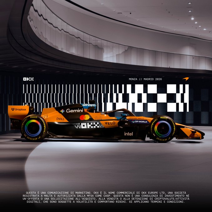 An orange and black McLaren Formula 1 race car displayed in a modern venue with 'OKX', 'MONZA // MADRID 2026', and various sponsor logos including Dropbox, Dell, Mastercard, and Velo visible.