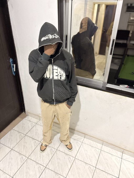 A person wearing a black hoodie with the word "AUTESIDE" and tan pants stands in front of a window, reflecting an image of themselves. The image's mysterious tone aligns with the post's cryptic message, "gm legends i pray you get unpaused & paid."