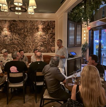 A group of people sit around a table at a dimly lit venue with decorative wallpaper and a chandelier, listening to a man standing and speaking.
