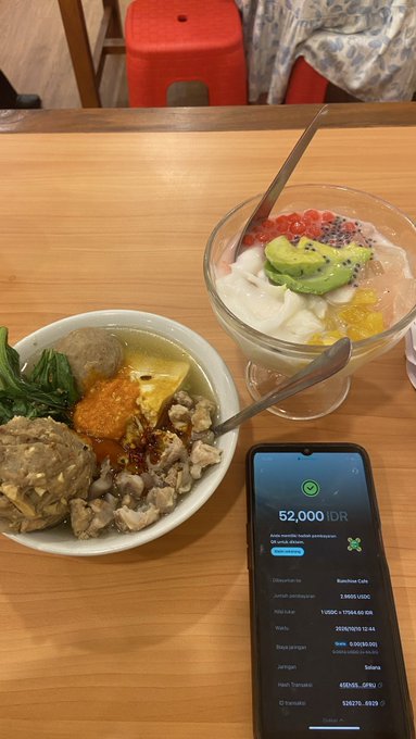 Two bowls of food sit on a wooden table alongside a smartphone displaying a balance of "52,000 IDR," with the phone screen showing various cryptocurrency conversions like Bitcoin and BNB.