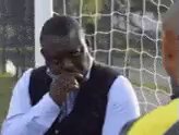 The image shows a man wearing a black vest, sitting down with his head resting on his hand. He appears to be deep in thought or contemplation. In the background, there are two other people present - one person standing behind him and another person sitting next to him. The scene seems to be taking place outdoors, possibly at a park or an open space where people gather for various activities.