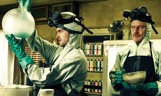 Two men in hazmat suits and gas masks handle chemicals in a makeshift laboratory, with one examining a flask of blue liquid and the other holding a bowl.