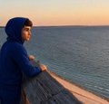 The image features a young boy wearing a blue hoodie, standing near a wooden railing overlooking the ocean at sunset. The boy appears to be looking out over the water with a thoughtful expression. In the background of the photo, there are two other people who seem to be enjoying their time as well. They appear to be in the distance and not directly involved with the main subject.