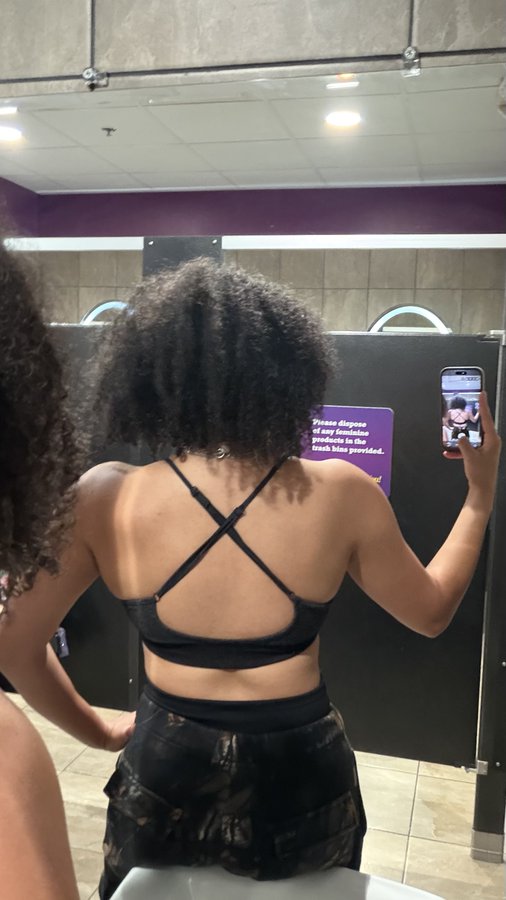 A woman with curly hair takes a mirror selfie in a public restroom, wearing a black sports bra with crisscross straps; a purple sign behind her reminds customers not to flush feminine products.