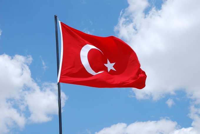 The image depicts a red Turkish flag flying high in the sky, with white clouds surrounding it. The photo is taken from a low angle, giving the impression of the flag fluttering in the wind against a bright blue sky. There is no text present in the image.