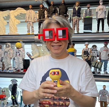 A smiling person wearing pixelated red square sunglasses holds a small yellow figurine, standing in front of shelves displaying various designer art toys and collectibles, connecting to the @nounsdao proposal by showcasing creator collaboration potential in the NFT collectibles space.