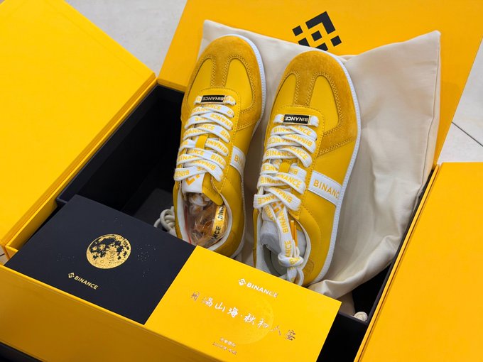 An open yellow Binance branded shoebox contains a pair of yellow sneakers with 'BINANCE' branding on the laces. A black box with a gold globe logo and Chinese characters sits inside alongside the shoes.