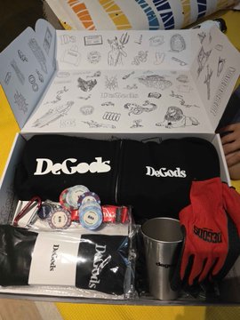 The image features a box containing various merchandise items related to the DeGodsNFT brand. The box contains a hoodie, gloves, and other accessories such as a cup and a pen. There are also some stickers inside the box that read "DeGods" and "Joined @DeGodsNFT at NFT Paris back in 2003. And never left since then. More than that, in the years that come, I manage to acquire my desire Ape Trait." The post seems to be promoting a merchandise collection from DeGodsNFT, which includes items like hoodies and gloves for fans of the DeGodsNFT brand.