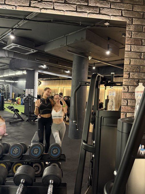 Two women in athletic wear pose together at a gym, holding dumbbells on a rack labeled "32" and "24," with industrial-style brick and metal decor behind them. The image reflects the post's "gm" (good morning) greeting to the Web3 community, suggesting the sender is starting their day with a workout session.