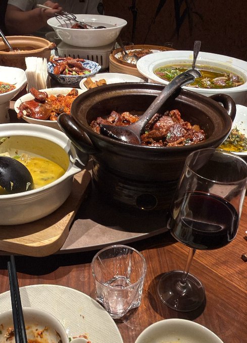 A dining table is covered with various Chinese dishes, including a clay pot of braised meat, soup, and other shared plates, alongside a glass of red wine and an empty glass. The image shows the casual setting where a dinner conversation about diversifying from $DOGE into $SPCX, both connected to Musk, is taking place.