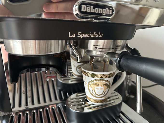 A close-up of a 'DeLonghi La Specialista' espresso machine pouring coffee into a Versace-branded cup decorated with the Medusa logo.