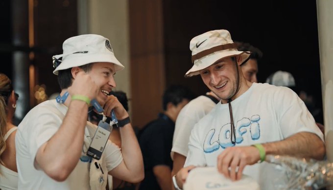 Two men wearing bucket hats (one Adidas, one Nike) and white t-shirts smiling and laughing at an outdoor event, with one holding a COFI-branded bag or item.