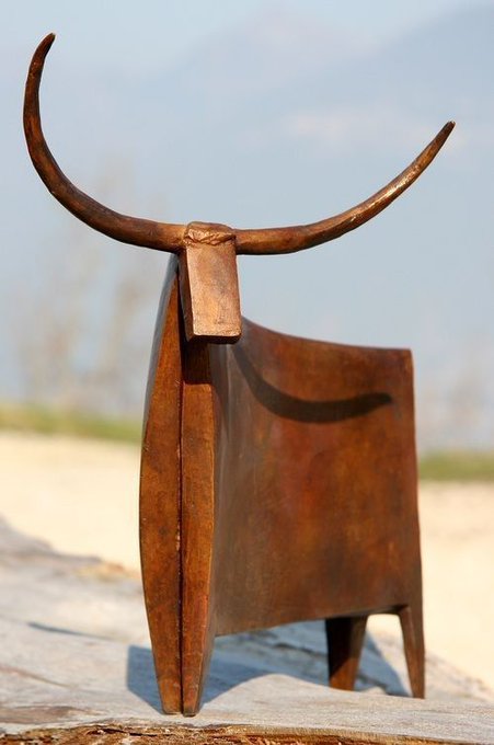 A rustic metal sculpture resembling a stylized bull with long curved horns stands on a wooden surface against a blurred sky background.