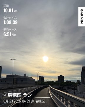 The image shows a city street with buildings, a bridge, and a sun setting over the horizon. The photo appears to have been taken during an evening run or walk, as indicated by the time stamp. The post features Japanese text that reads "朝ラン10キロろ ゆっくりジョグ 今週はロング走の予定だったけど雨予報 台風も来そうで予定が狂いますね,焦らそうに休みも入れて頑張ります". The image and the post are related to each other as they both provide information about an evening activity in a foreign language.