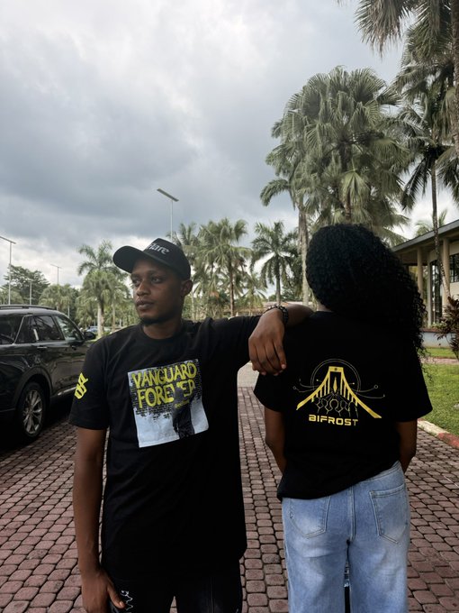 Two people wearing matching 'Vanguard Forest / Bifrost' black t-shirts posing on a brick path with palm trees in the background.
