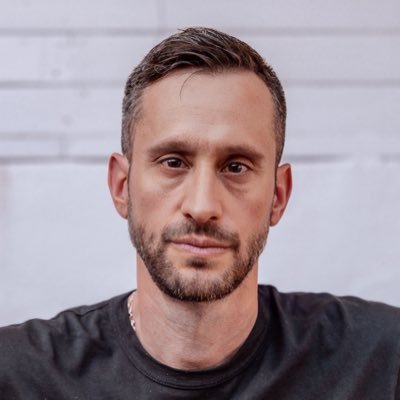 The image features a man with a beard wearing a black shirt, looking directly at the camera. The background of the photo consists of white walls, which contrasts with the man's dark clothing. There are no texts or other objects present in the image, making it a simple and uncluttered composition that highlights the subject - the bearded man in the black shirt.