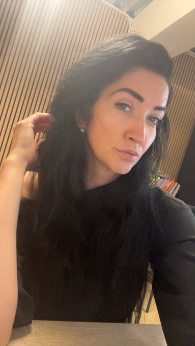 A woman with long dark hair takes a selfie, wearing a black top and small earrings, with a slatted wood wall and a shelf of colorful books in the background. The image conveys a relaxed Friday mood, pairing with her "GM everyone! TGIF" message to greet followers and celebrate the weekend ahead.