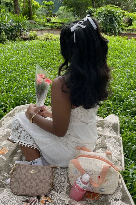 A young woman with long dark hair, a white bow, and a white lace dress sits on a blanket holding pink carnations, with a Coach crossbody bag, a yellow checkered pet-themed bag, and a pink drink nearby, evoking the relaxed, welcoming vibe of a "gm" greeting. The serene garden setting complements the friendly "let's connect" message, suggesting an inviting and stylish morning moment.