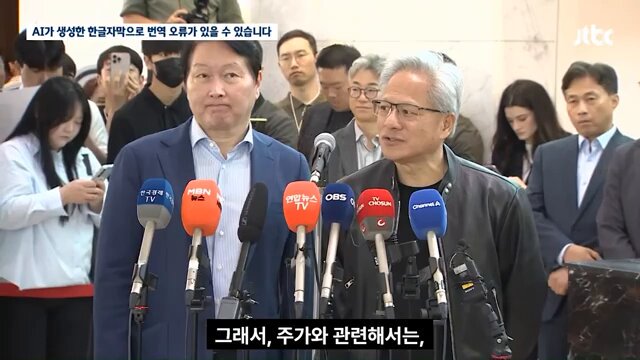 The image shows a man wearing a suit standing next to another man who is also dressed in a suit, both holding microphones. They are surrounded by several other people, some of whom appear to be taking pictures or recording videos with their cameras. The background reveals that they are at an event where multiple people are present. The focus on the two men and their microphones suggests that they might be speaking or addressing an audience during this gathering.