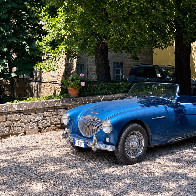 A classic blue convertible sports car is parked on a gravel courtyard beside stone walls, flowers, and mature trees.