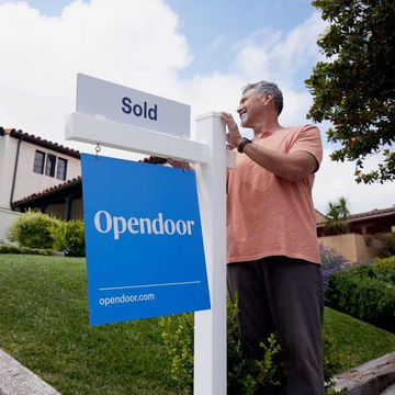 A smiling older man installing an Opendoor 'Sold' real estate sign in front of a suburban home.