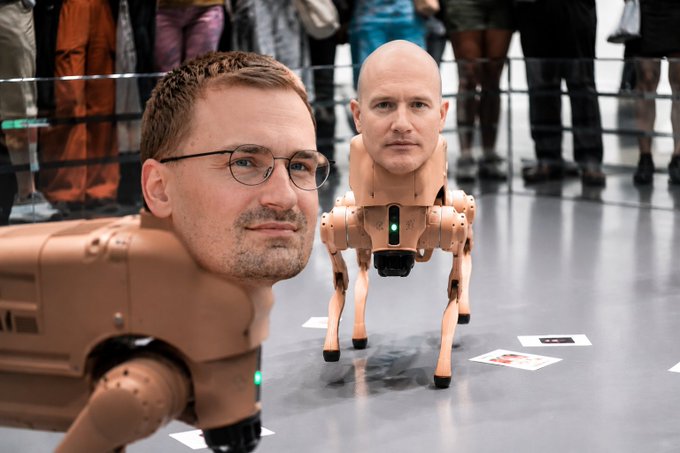 A man wearing glasses poses in the foreground next to a humanoid robot with a hyper-realistic human head and a beige mechanical body on four legs, displayed at what appears to be an exhibition.

The image seems unrelated to the post's criticism of Coinbase's Brian Armstrong, instead depicting an unrelated tech exhibition featuring a humanoid robot.