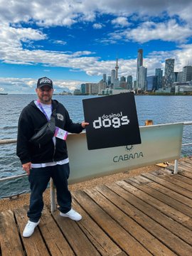A man wearing a 'STAY' cap and black jacket stands on a wooden deck holding a black 'Copool dogs' sign, with a city skyline visible across the water behind him.