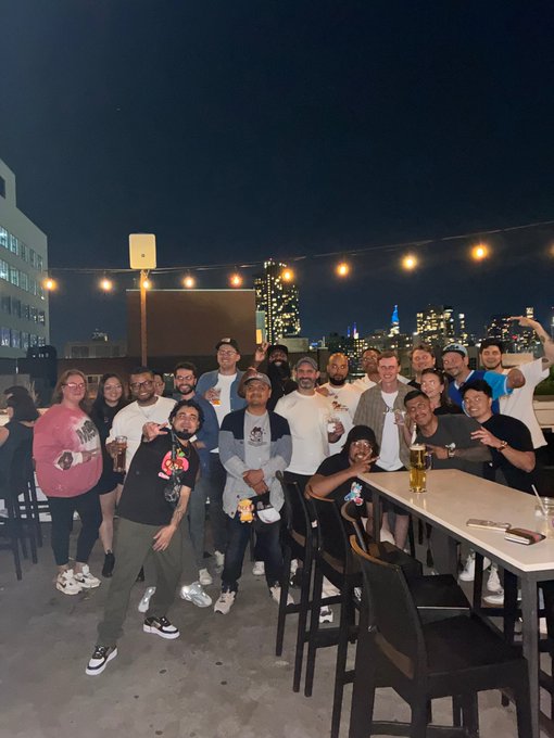 A large group of people poses for a nighttime rooftop gathering with string lights overhead and a lit city skyline in the background.
