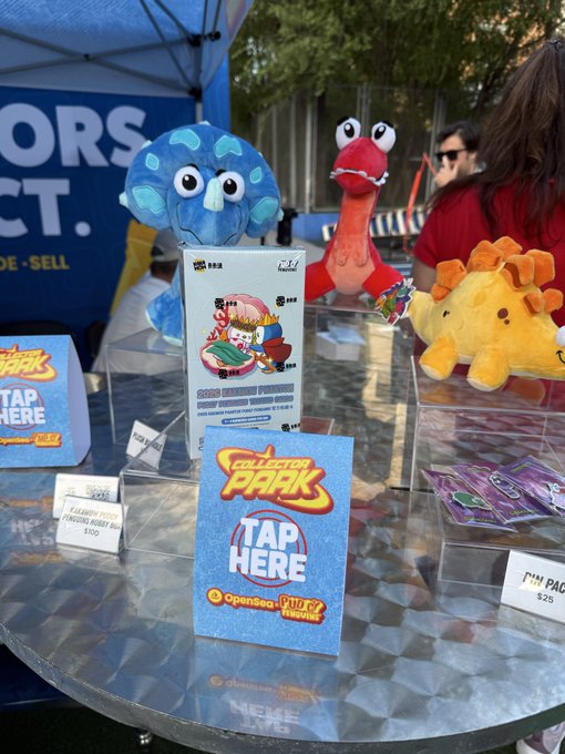 A vendor booth displaying 'Collector Park' merchandise, including plush toys and trading cards, with signs reading 'TAP HERE' and 'OpenSea' and prices like '$100' and '$25'.