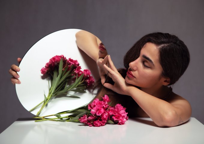A woman with dark hair poses elegantly beside a round mirror and a bouquet of vibrant pink flowers on a white surface.