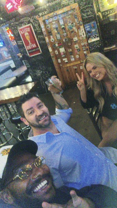 A selfie of Adam Levine smiling in a bar, accompanied by two other people, with a 'Yahtzee' sign visible in the background.