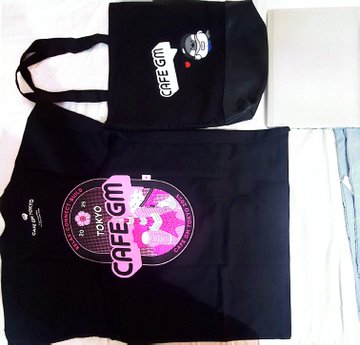 Two black "Cafe GM" tote bags, one featuring a pink vinyl record graphic labeled "ROMO 2" and the other displaying the Cafe GM logo, are laid out on a bed as a promotional package gifted to a content creator. The image complements the post's space-themed announcement by showcasing the merchandise sent as a thank-you for the collaboration.