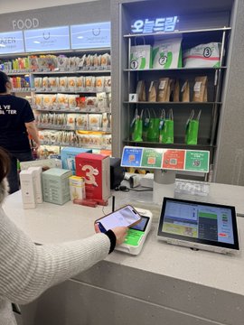 A convenience store checkout counter where a person uses a smartphone on a reader, with Korean text '오늘의텀' visible on a refrigerated display in the back.