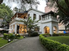 A photograph of an elegant, multi-story classical-style mansion with arched entryways and tall windows, set behind a gated driveway with manicured hedges and trees.