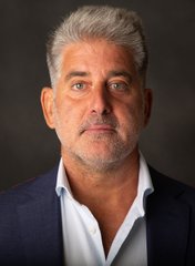 A professional headshot portrait of a middle-aged man with silver hair wearing a blue blazer over a light blue shirt.