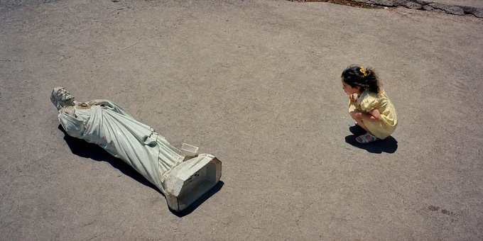 A young girl in a yellow dress crouches on cracked pavement to look closely at a toppled stone statue lying on the ground.