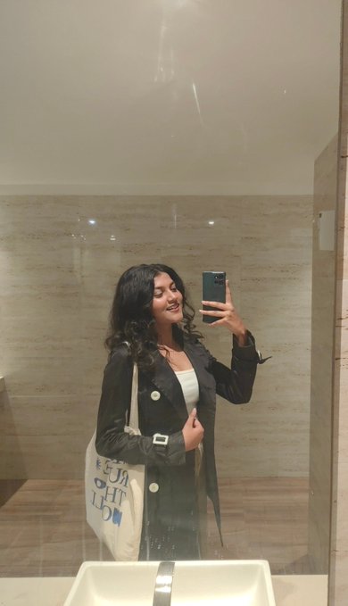 A young woman with long dark hair takes a mirror selfie in a modern bathroom, wearing a black jacket and white top while holding up her phone to capture the moment, her cream tote bag reading "HUG LIFE" visible beside her as she smiles in celebration of her impressive milestone of 50k+ impressions and two finalized projects.