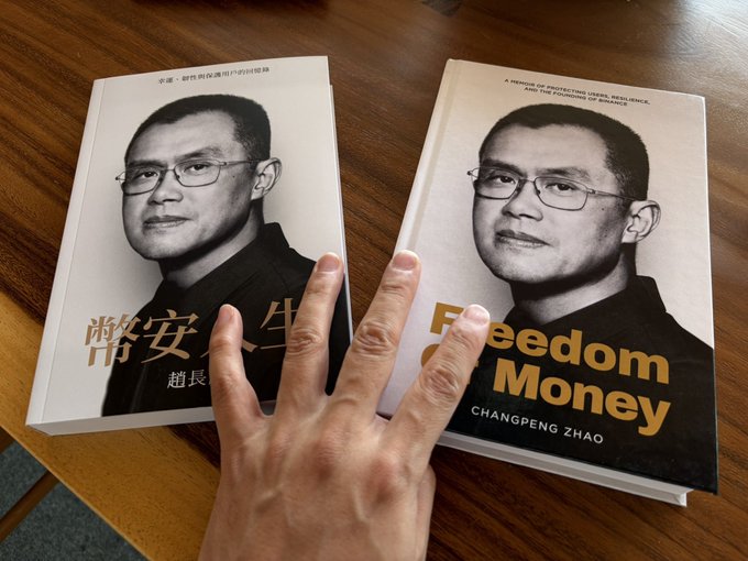 Two copies of Changpeng Zhao's memoir "Freedom Money" lie on a wooden table—one in English and one in Chinese (幣安人生)—being displayed by a hand reaching toward them, likely photographed to show support for CZ ahead of his appearance in Hong Kong.
