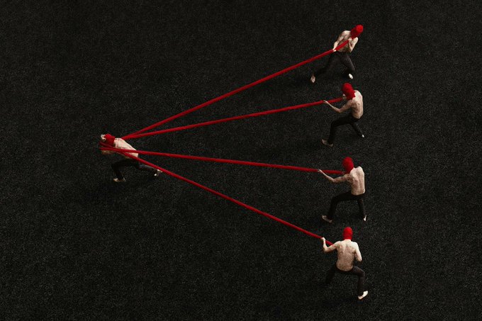 An artistic top-down shot of people in white shirts and red caps pulling red ropes connected to another person on dark grass, creating a dynamic visual composition.
