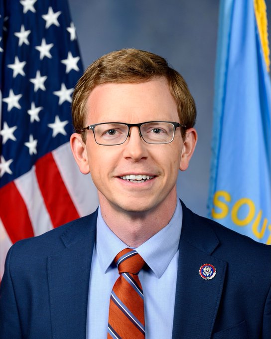 The image features a man wearing glasses, a blue suit jacket with a red tie, and an American flag behind him. The man appears to be smiling for the camera while posing against this backdrop. The caption of the image reads "Let'S get it done! #metallicus #XPRNetwork #metalblockchain #webauthwallet #loanprotocol #MTL". This caption suggests that the man is likely a professional or someone involved in the financial industry, given his attire and the presence of an American flag.