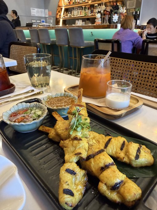 A photo of a meal at a bar or restaurant featuring grilled chicken satay skewers on a black plate, accompanied by dipping sauces, an orange drink, and water.