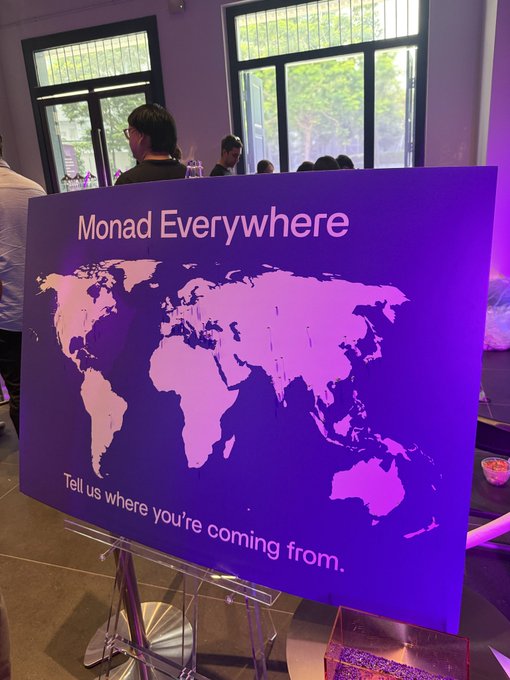 A purple sign displays a world map with the text "Monad Everywhere" and "Tell us where you're coming from.", set up indoors near windows.