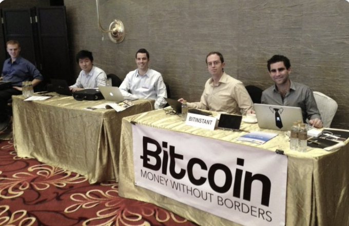 Five men sit at a table draped with a 'Bitcoin Money Without Borders' banner, with a 'BitInstant' sign visible, working on laptops during what appears to be a Bitcoin-related event.