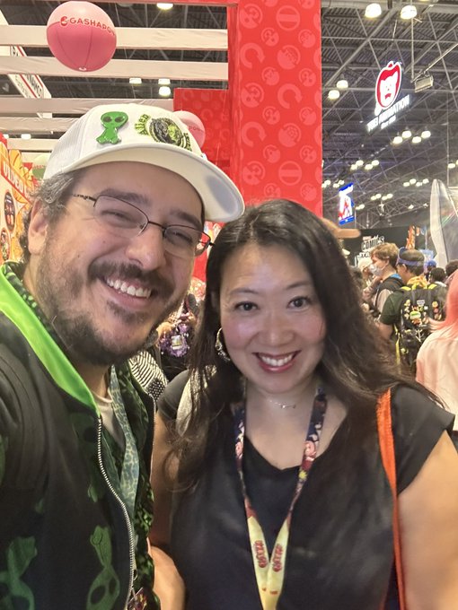 A man with glasses and a beard wearing a white cap and green scarf poses for a selfie with a smiling woman wearing a lanyard, in what appears to be a convention hall with a red 'GASHADOKURO' balloon visible above.