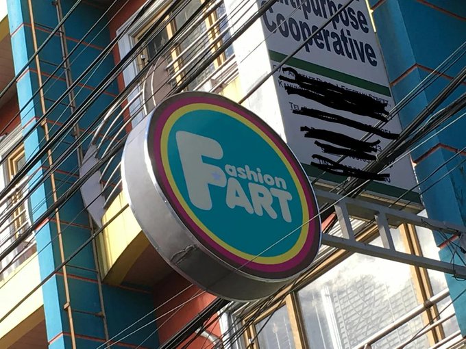 A storefront sign reading 'Fashion FART' hangs outside a multi-story building with many electrical wires crossing in front of it.