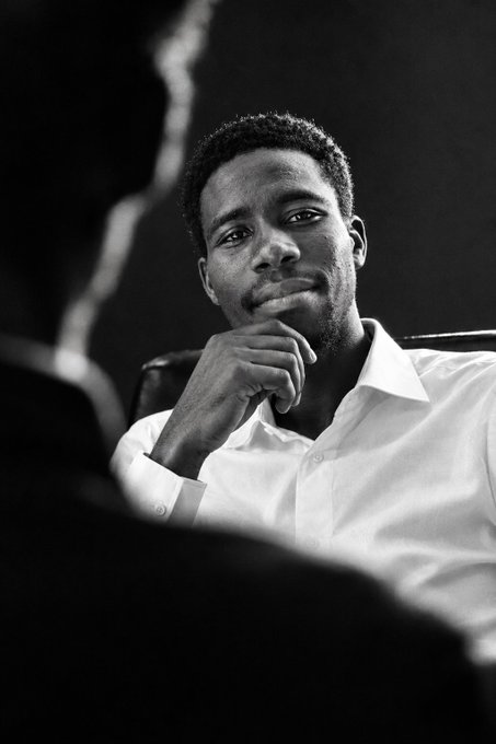 A black and white photograph of a young man wearing a white shirt, resting his chin on his hand while looking thoughtfully toward the camera.