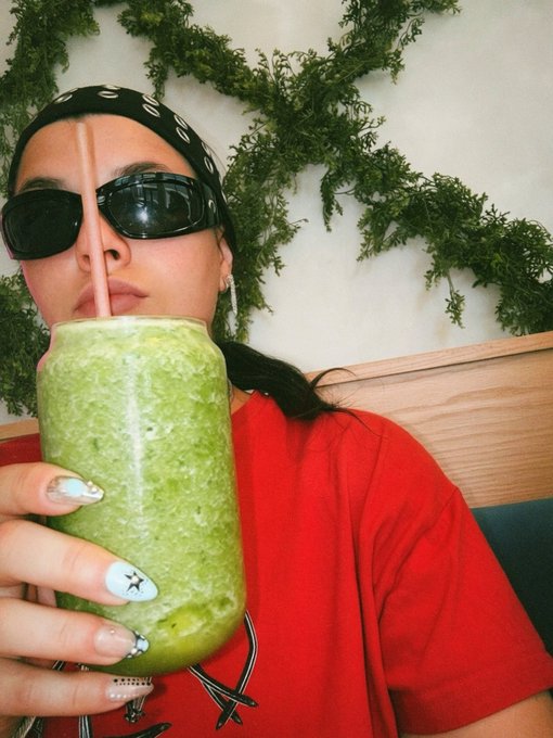 A woman wearing a black bandana, dark sunglasses, and a red shirt drinks a green smoothie from a glass jar through a straw.