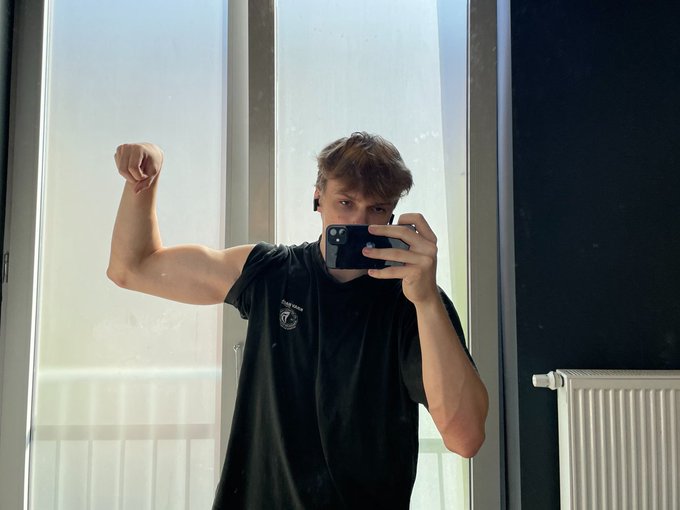 A young man wearing a black t-shirt and earbuds takes a mirror selfie with his phone while flexing his right bicep, standing in front of a bright window with a radiator visible to the right.