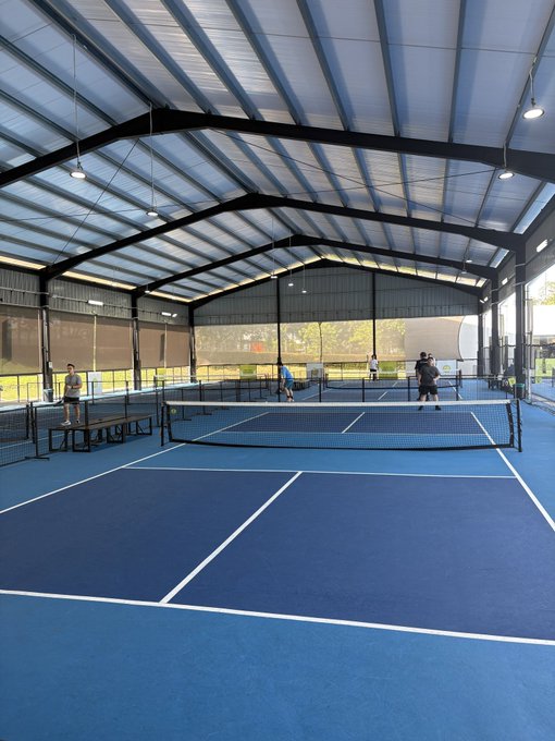 An indoor blue tennis and pickleball facility with a high arched roof, where several people are playing on divided courts with nets under bright natural lighting.